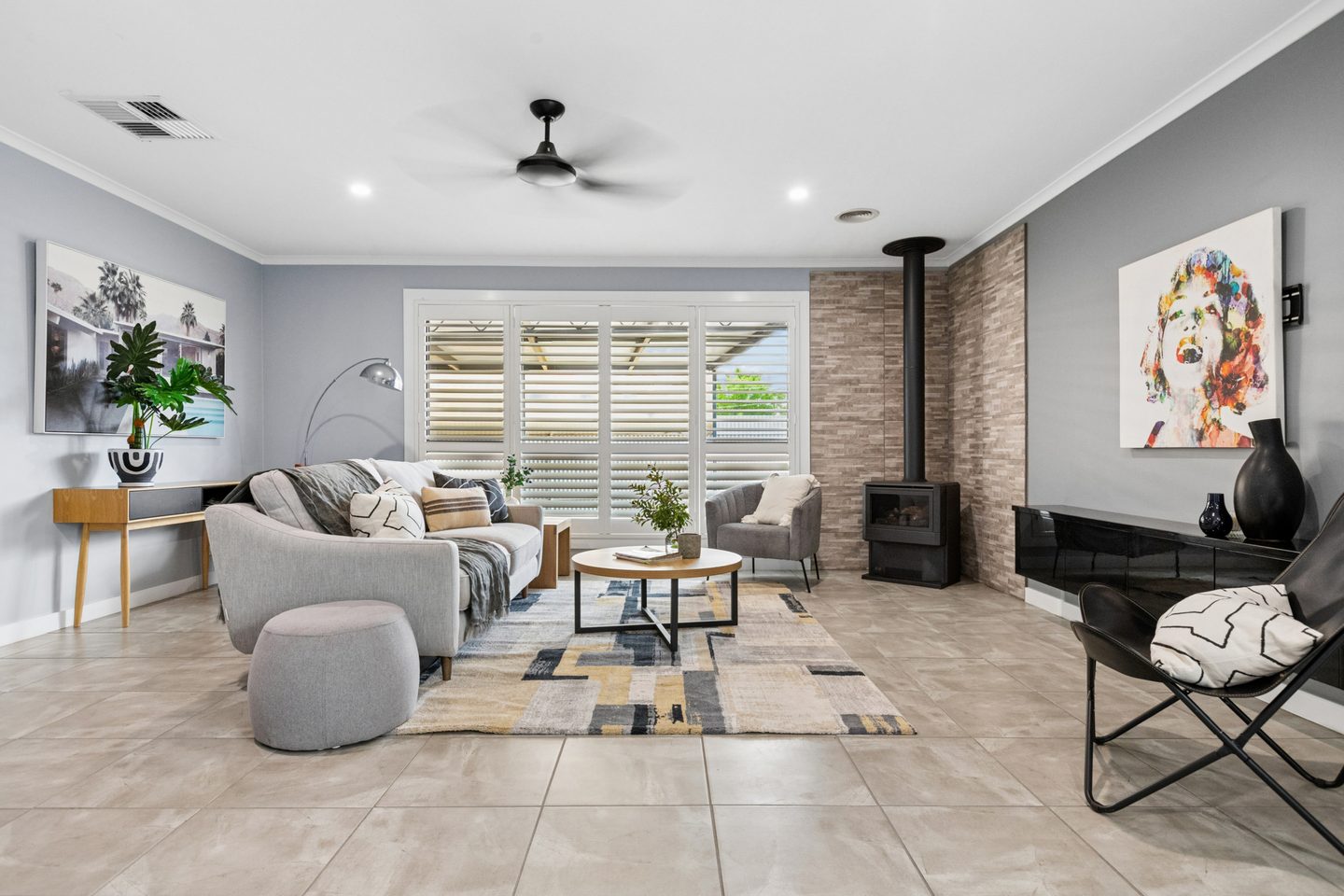 Living room with a wood heater, white shutters, tiled floor and ceiling fan